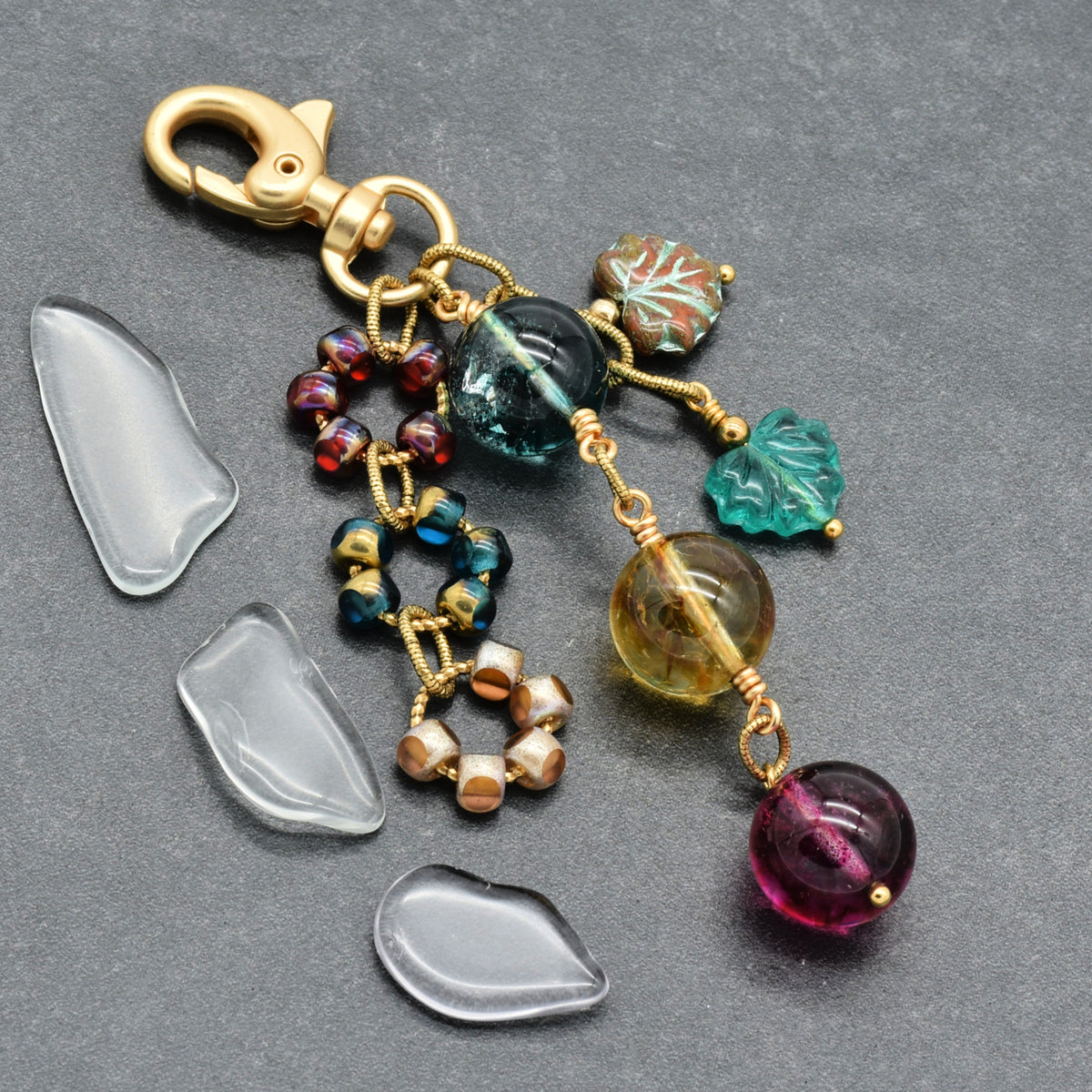 A gold-toned keychain or bag charm with multiple dangling beads and charms, including a large dark blue-green round bead, a translucent yellow-green bead, a purple round bead, leaf-shaped charms, and a cluster of iridescent beads. Three clear teardrop-shaped embellishments are placed beside it on a dark gray textured background.