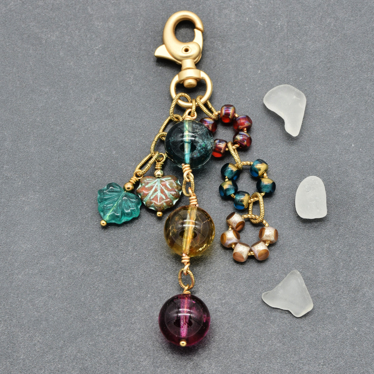 A handmade beaded keychain with a gold-toned lobster clasp, featuring multiple charms including a green leaf, a patterned bead, a dark blue bead, a yellow-brown bead, a gold ring, and a large purple round bead, displayed on a gray background with four small translucent stone chips.