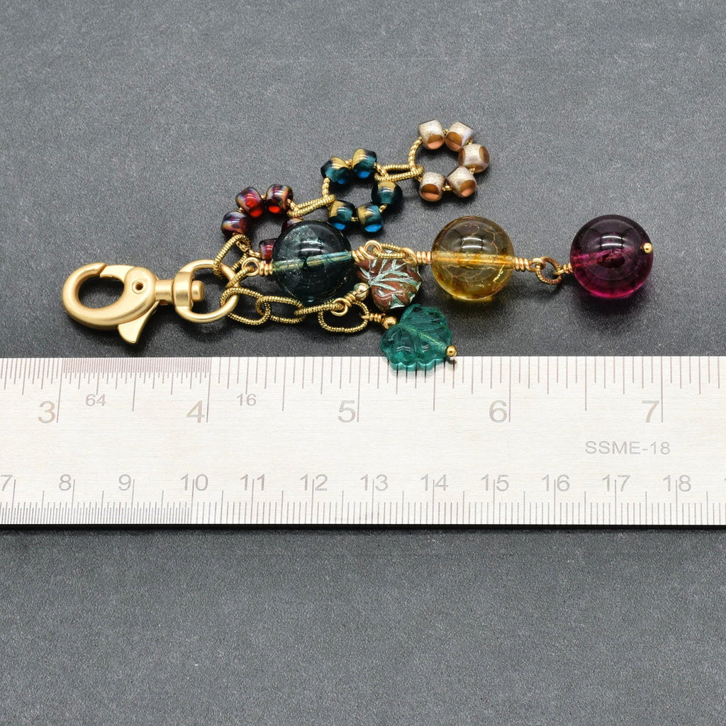 A handmade beaded keychain or bag charm with a gold-tone lobster clasp, featuring various colorful glass beads and crystal charms, shown next to a metal ruler for scale on a dark gray background.