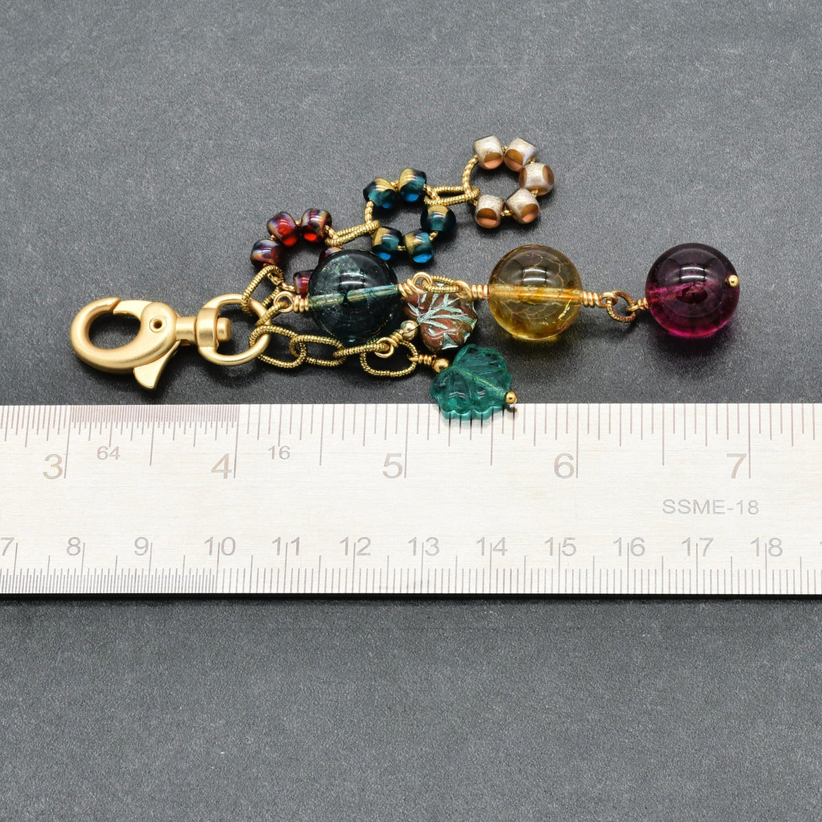 A handmade beaded keychain or bag charm with a gold-tone lobster clasp, featuring various colorful glass beads and crystal charms, shown next to a metal ruler for scale on a dark gray background.