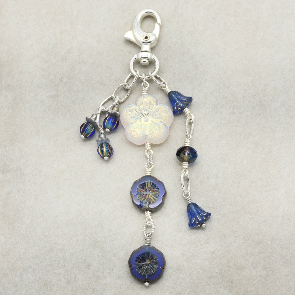 A silver-toned lobster clasp keychain or bag charm featuring a white iridescent flower bead, blue faceted beads, and small blue bell-shaped beads, displayed on a neutral light gray background.
