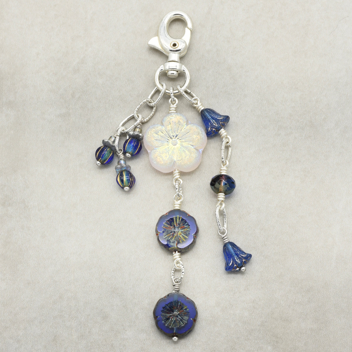 A silver-toned lobster clasp keychain or bag charm featuring a white iridescent flower bead, blue faceted beads, and small blue bell-shaped beads, displayed on a neutral light gray background.