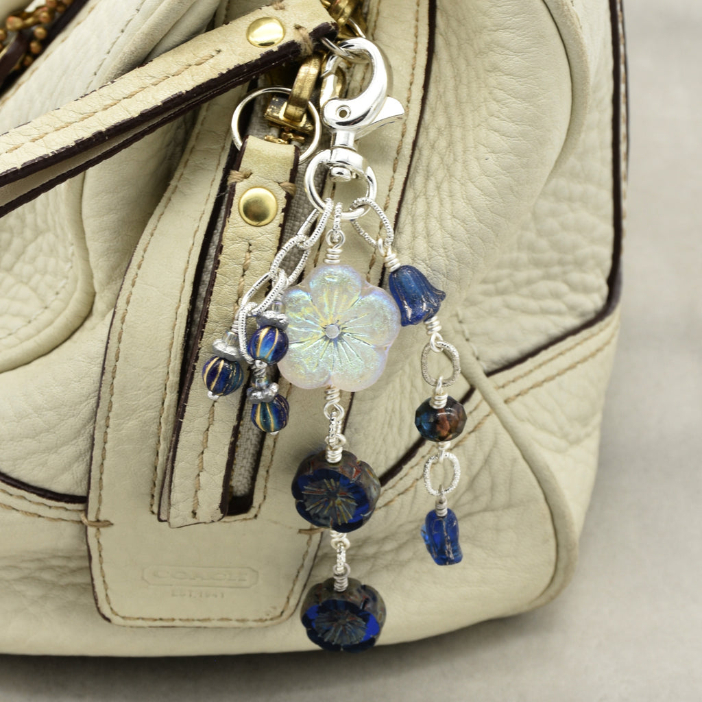 A close-up of a handbag in light beige textured leather with a silver-tone keychain charm featuring a white iridescent flower and dark blue faceted beads.