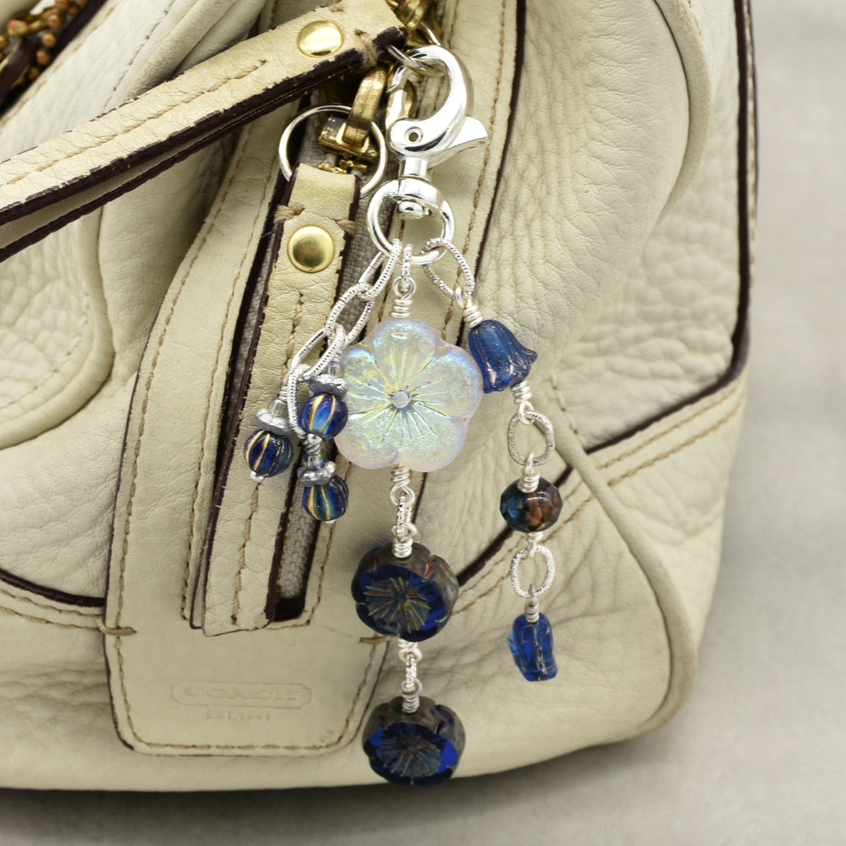 A close-up of a handbag in light beige textured leather with a silver-tone keychain charm featuring a white iridescent flower and dark blue faceted beads.