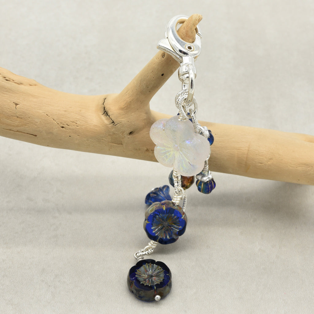 A close-up of a silver-toned clip-on earring with dangling blue glass beads and a milky white flower charm, displayed on a light wood branch against a neutral gray background.