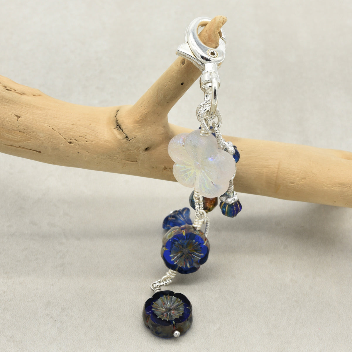 A close-up of a silver-toned clip-on earring with dangling blue glass beads and a milky white flower charm, displayed on a light wood branch against a neutral gray background.