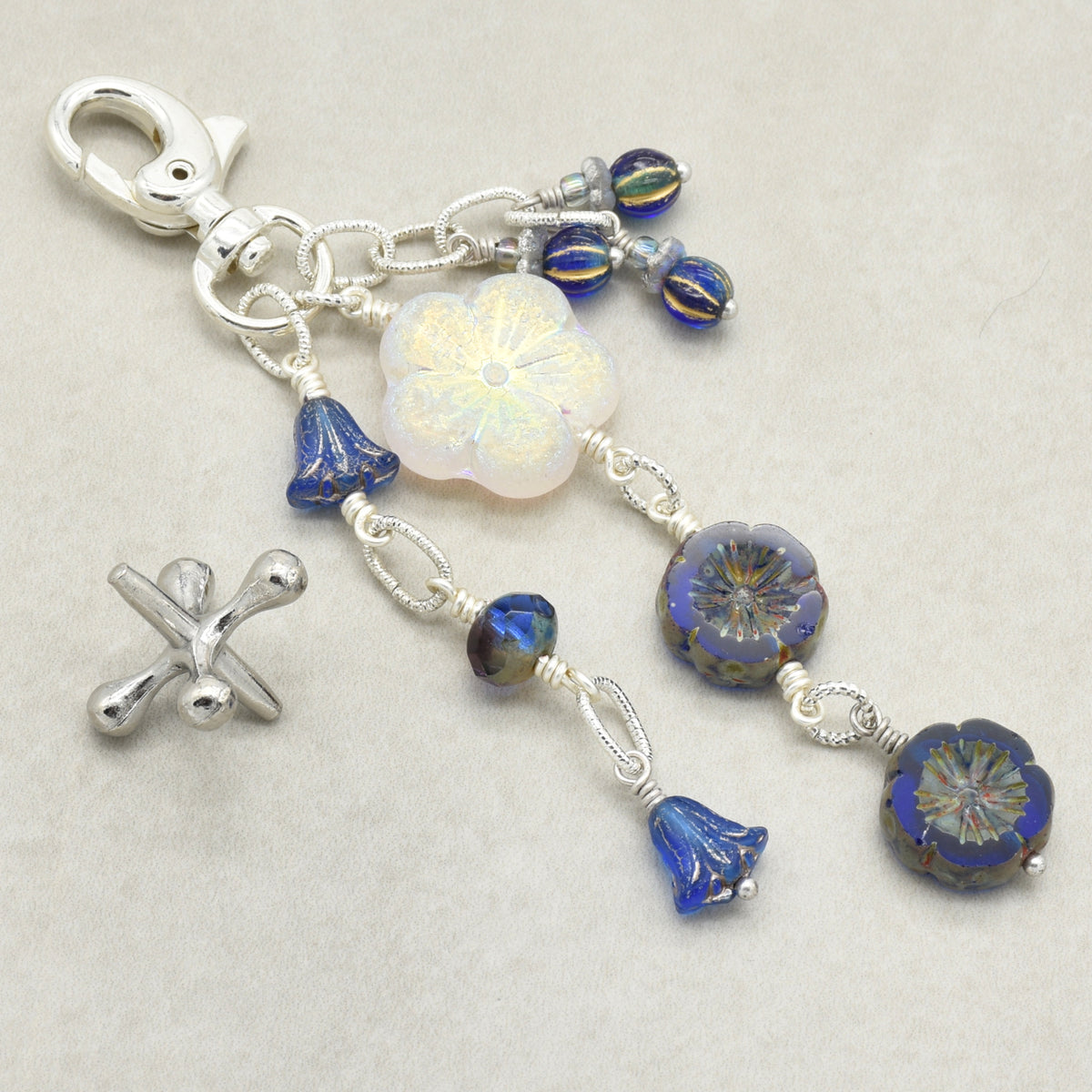A handmade beaded keychain featuring a silver lobster clasp, a white iridescent flower bead, blue glass bell beads, patterned blue and gold flower beads, displayed on a neutral beige background.