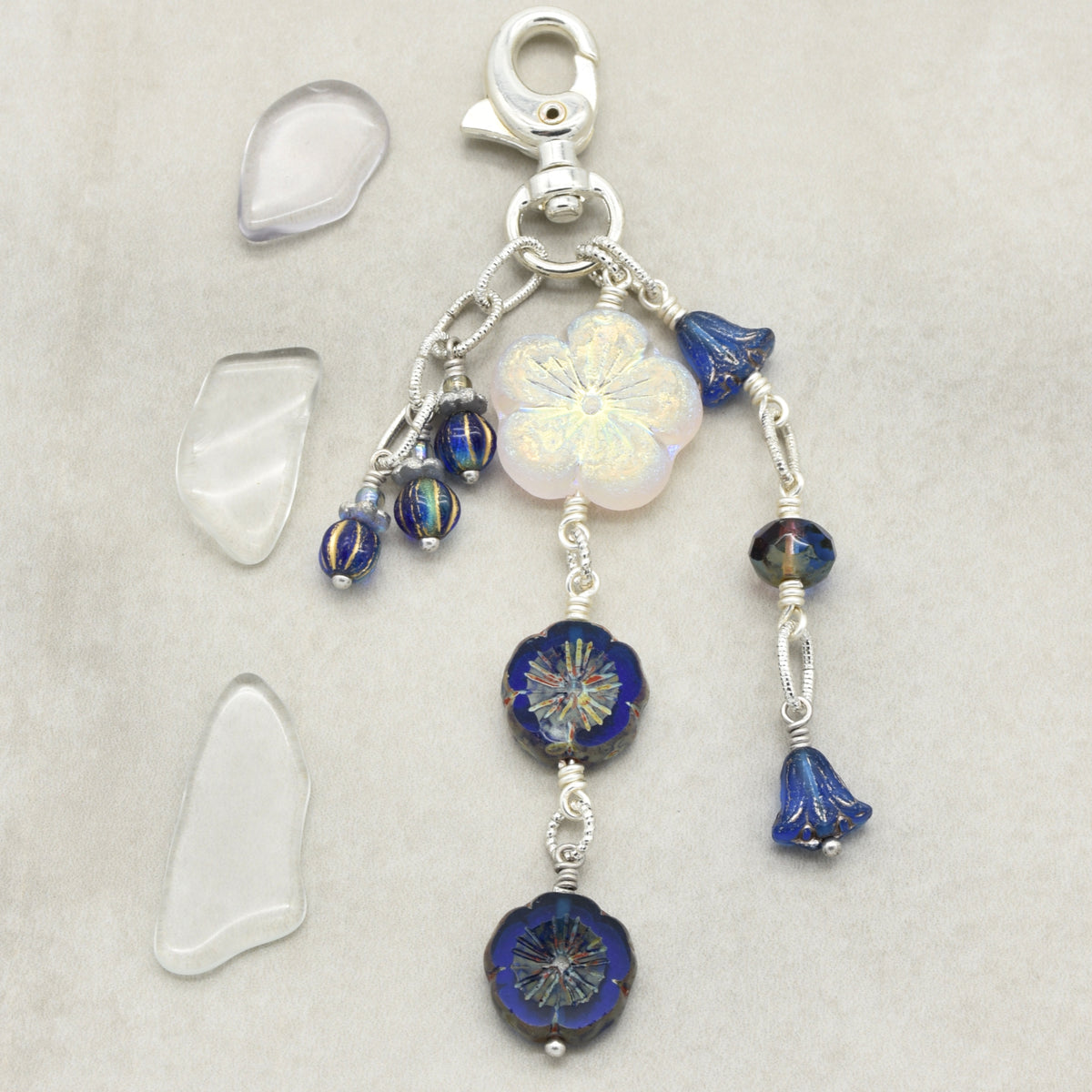 A silver-toned beaded charm with a lobster clasp, featuring a white iridescent flower, and deep blue faceted beads, displayed on a neutral beige background.