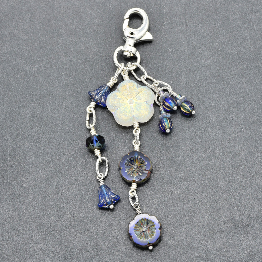 A silver-toned beaded charm keychain featuring a white flower, blue glass beads, and floral disc charms, displayed on a dark gray textured background.
