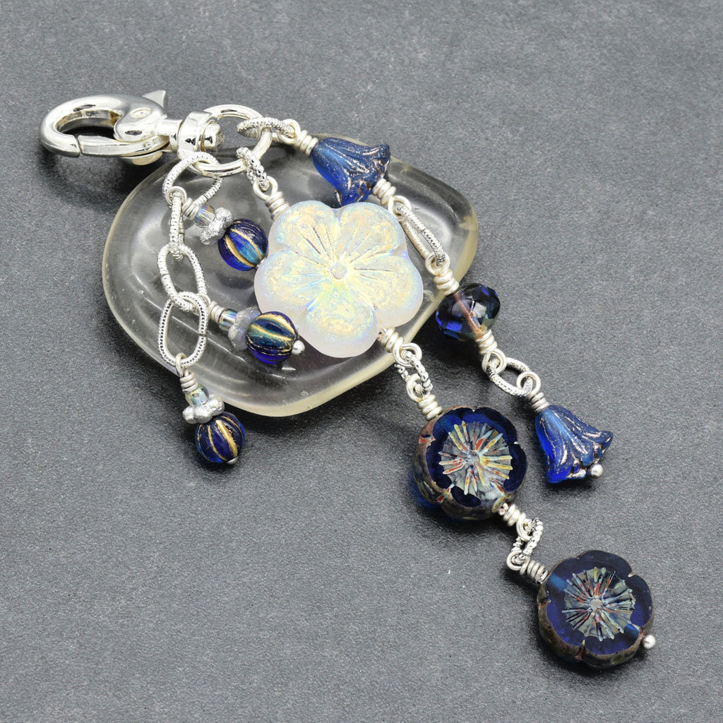 A handmade glass keychain or bag charm featuring a silver-tone clasp, a white iridescent flower bead, and multiple dangling blue glass beads and floral elements on silver wire, set against a dark gray textured background.