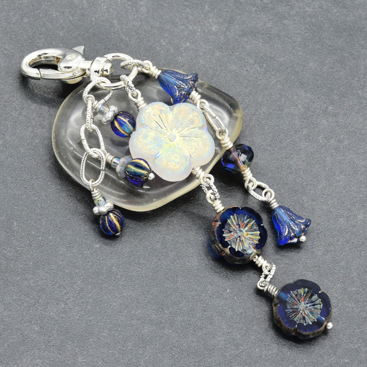 A handmade glass keychain or bag charm featuring a silver-tone clasp, a white iridescent flower bead, and multiple dangling blue glass beads and floral elements on silver wire, set against a dark gray textured background.