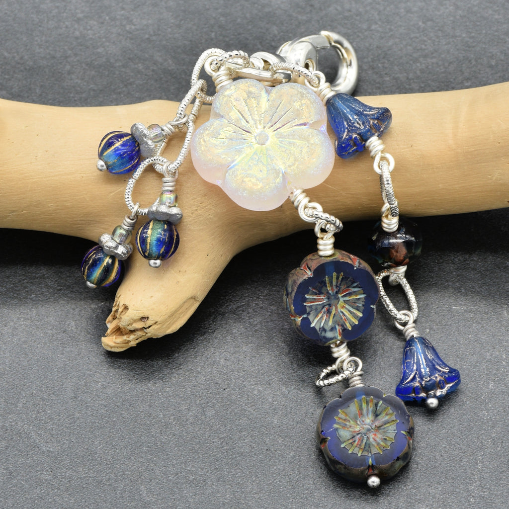 A handmade glass bead bracelet with a large iridescent white flower charm, blue bell beads, and patterned blue beads, displayed on a light wood branch against a dark gray background.