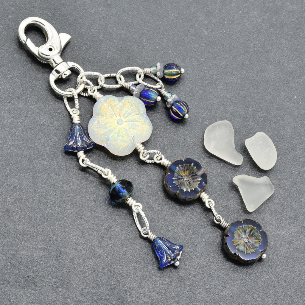 A silver-toned keychain or bag charm with a lobster clasp, featuring a large iridescent white flower bead, and blue faceted beads, displayed on a dark gray textured background.