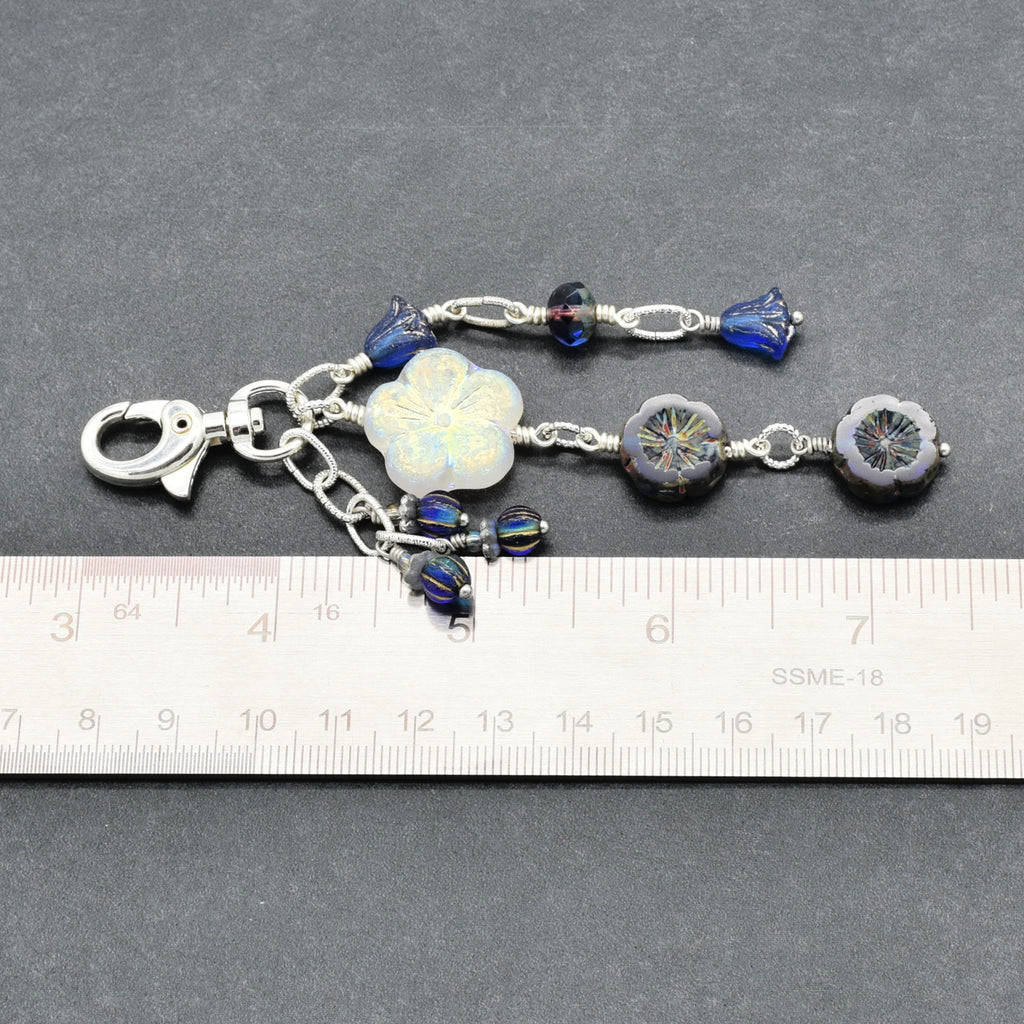 A silver-toned beaded keychain or bag charm with a lobster clasp, shown on a dark gray background with a ruler at the bottom for scale. The charm features a white iridescent flower bead, blue glass beads, and dark blue floral beads.