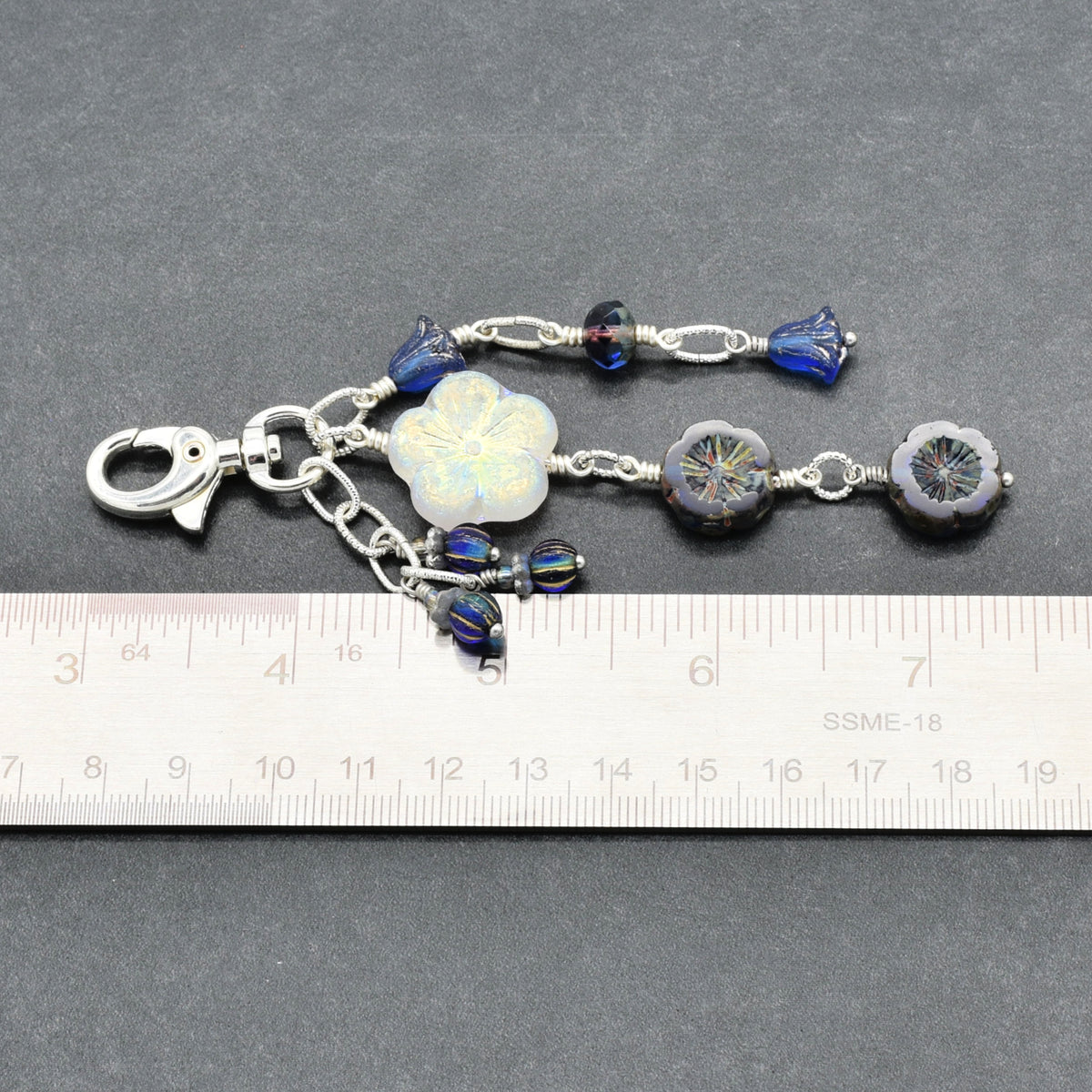 A silver-toned beaded keychain or bag charm with a lobster clasp, shown on a dark gray background with a ruler at the bottom for scale. The charm features a white iridescent flower bead, blue glass beads, and dark blue floral beads.