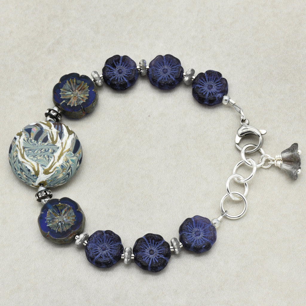 A beaded bracelet featuring dark blue glass beads with floral patterns, a large round patterned artisan polymer clay bead, and silver-toned metal links and charm on a neutral beige background.