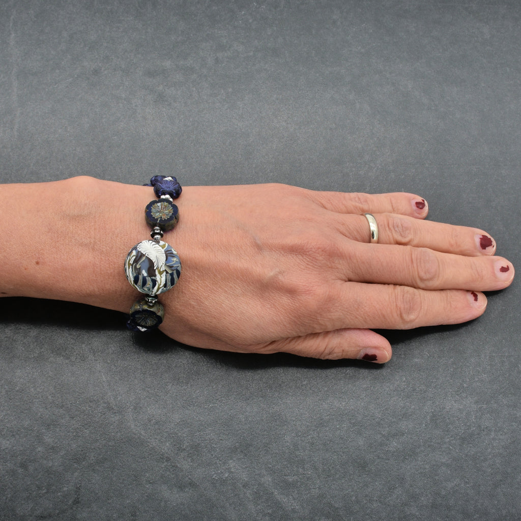 A beaded bracelet with dark blue, and silver-toned beads, including a large polymer clay bead, shown on a person's wrist against a dark gray textured background.