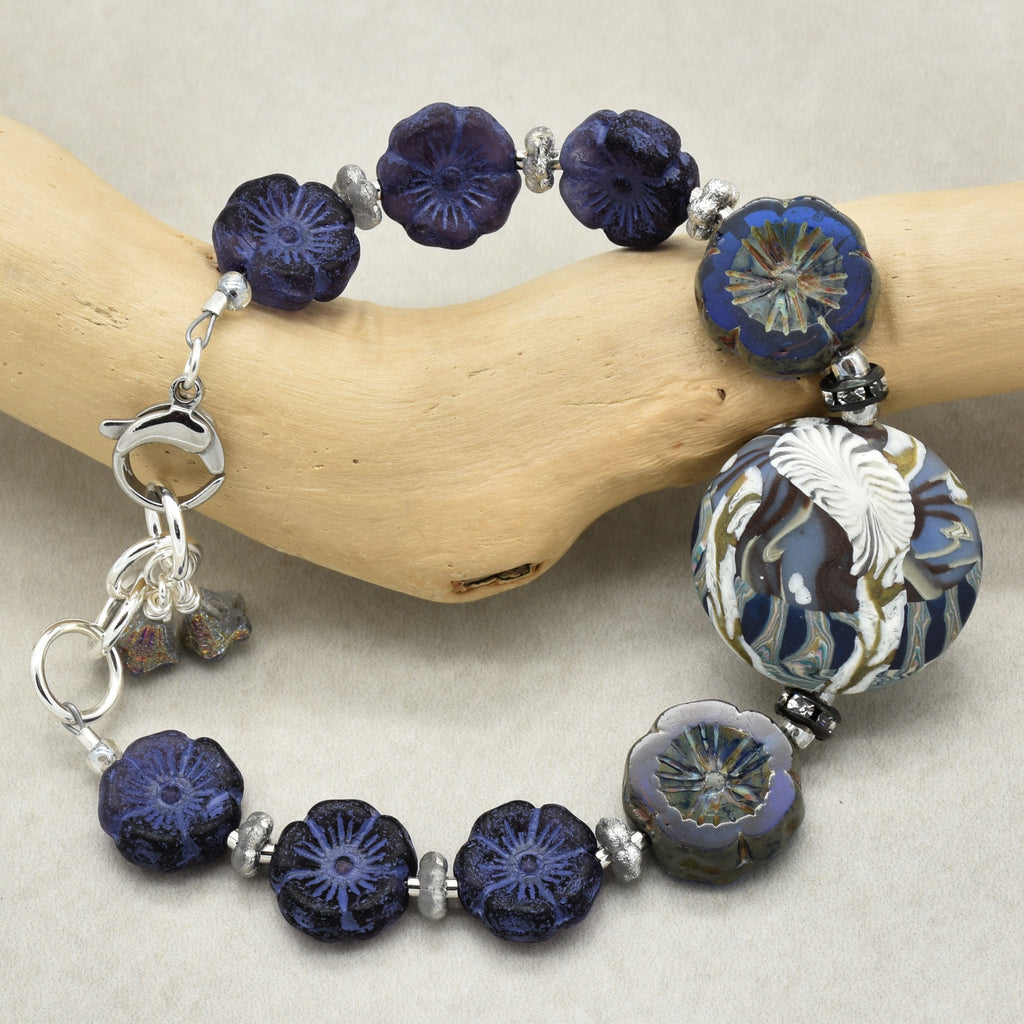 A beaded bracelet with dark blue and indigo flower-shaped beads, a large white and blue floral bead, and silver-toned spacer beads, displayed on a curved piece of natural wood against a neutral beige background.