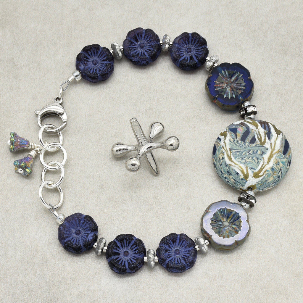 A beaded bracelet with dark blue patterned glass beads, a large round floral blue and gold bead, a silver clasp, and a small silver bellflower charms, displayed on a light beige background.