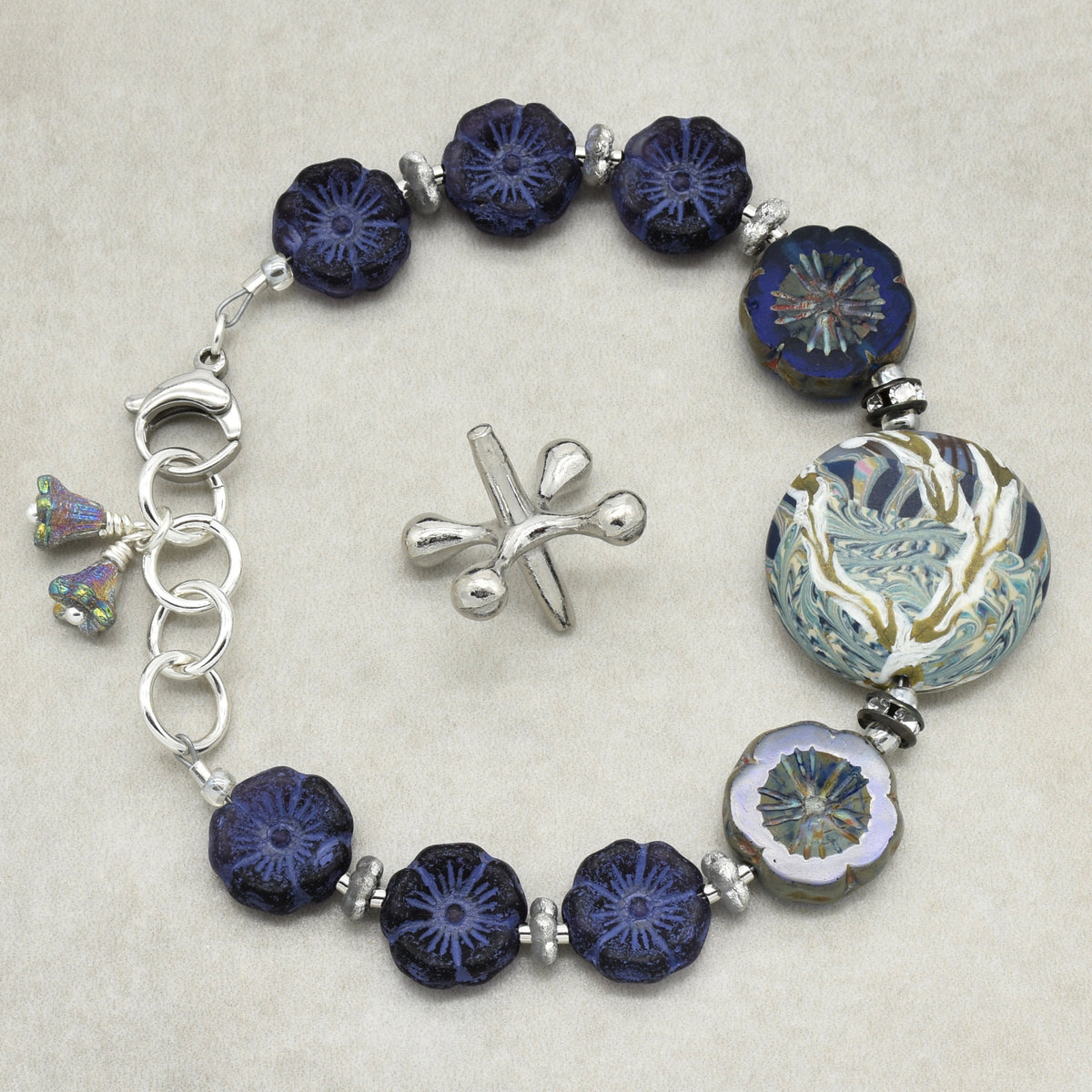 A beaded bracelet with dark blue patterned glass beads, a large round floral blue and gold bead, a silver clasp, and a small silver bellflower charms, displayed on a light beige background.