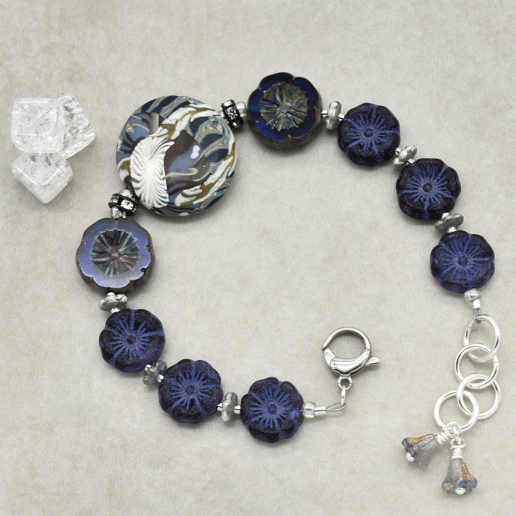 A beaded bracelet with dark blue and navy floral beads, a large patterned round bead, silver spacer beads, and silver-toned clasp and charm details, displayed on a neutral textured background.