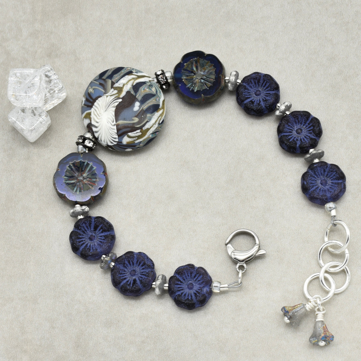 A beaded bracelet with dark blue and navy floral beads, a large patterned round bead, silver spacer beads, and silver-toned clasp and charm details, displayed on a neutral textured background.
