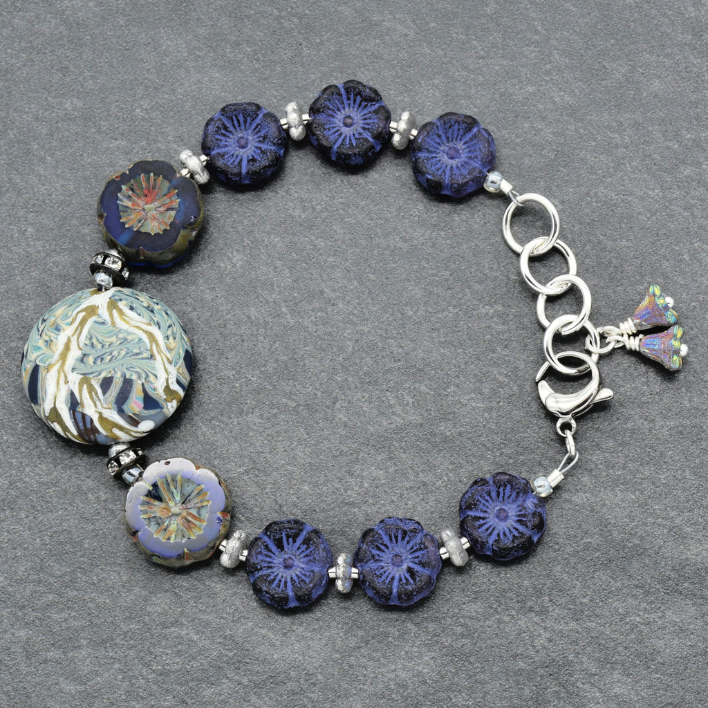 A beaded glass bracelet featuring dark blue and indigo floral beads, a large round blue and gold patterned bead, and silver-toned metal links and spacer beads, displayed on a textured gray background.