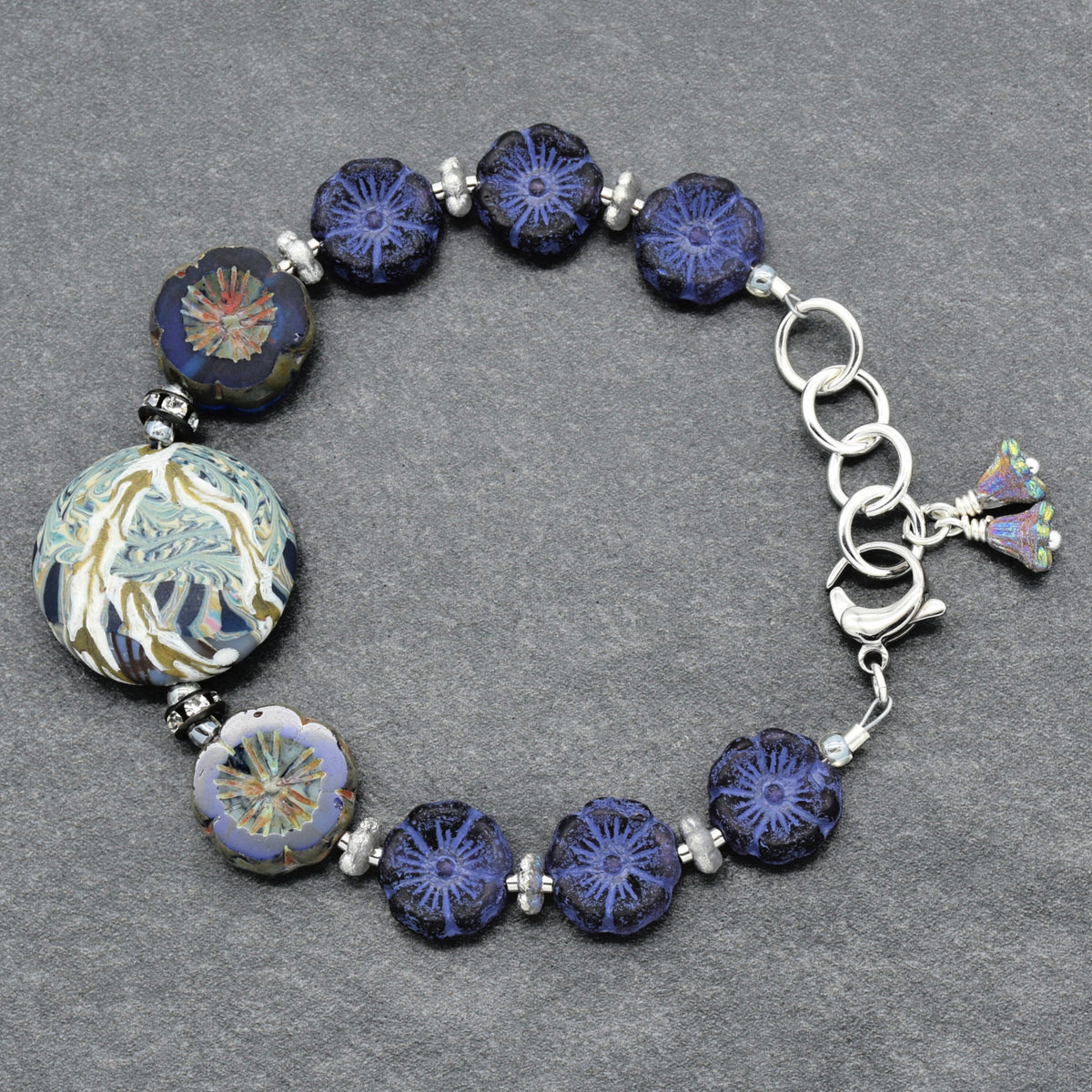 A beaded glass bracelet featuring dark blue and indigo floral beads, a large round blue and gold patterned bead, and silver-toned metal links and spacer beads, displayed on a textured gray background.