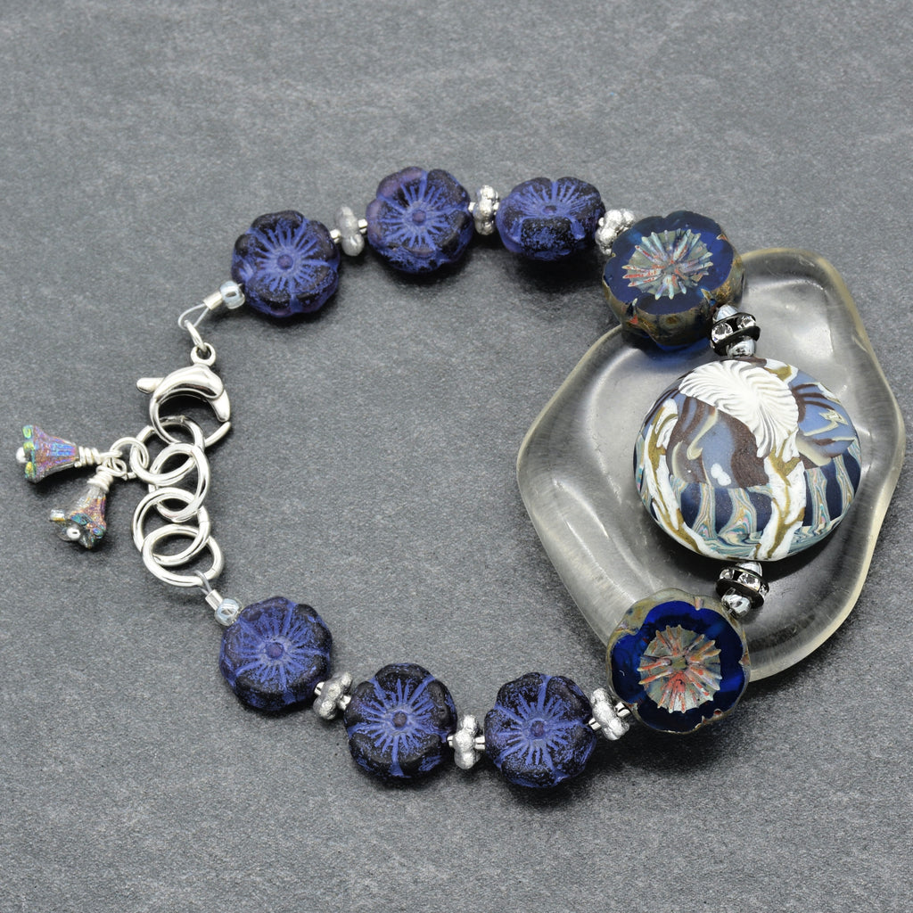 A beaded bracelet with dark blue and indigo floral glass beads, a large white and blue patterned bead, and silver-toned metal links and charm, displayed on a gray textured background with a clear stone prop.