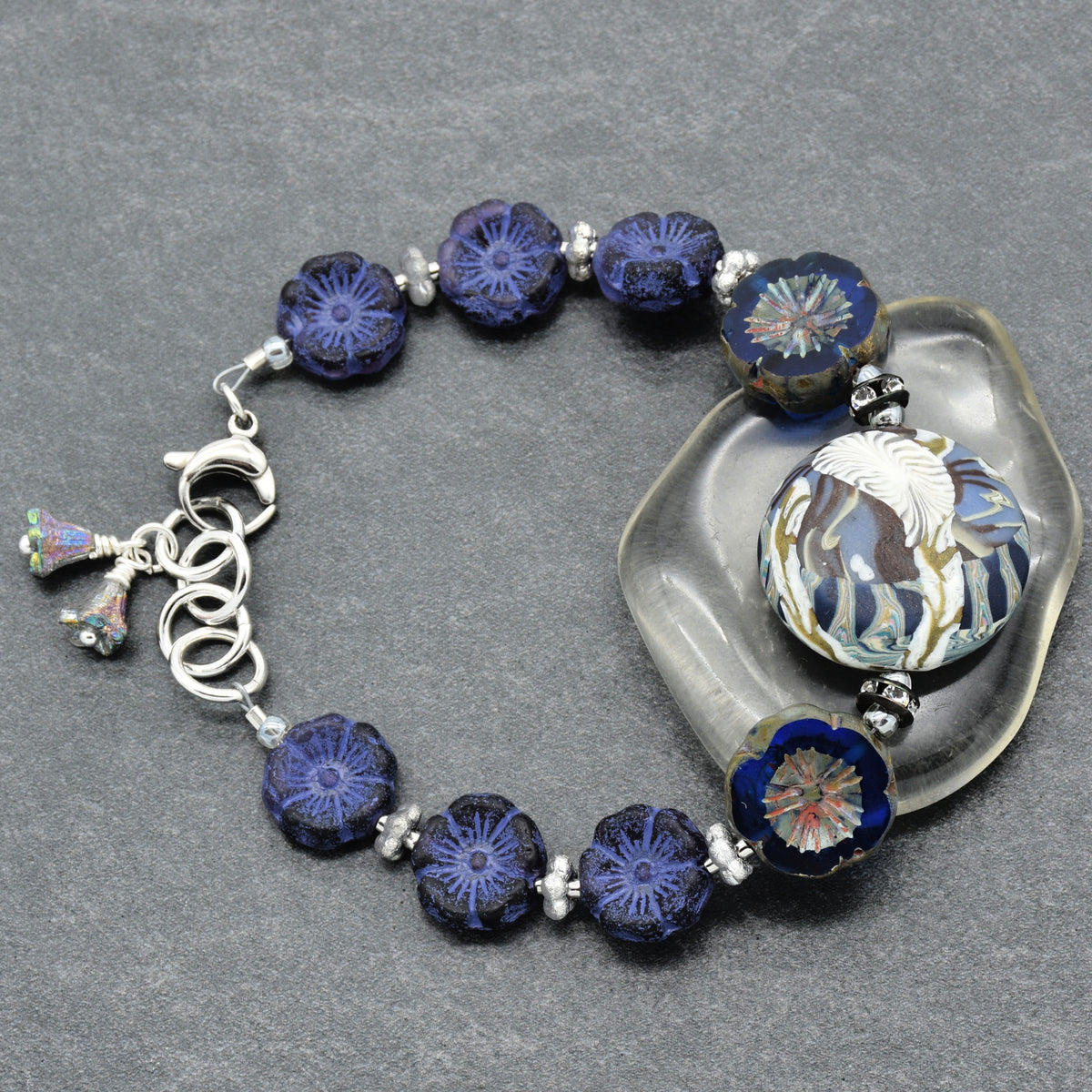 A beaded bracelet with dark blue and indigo floral glass beads, a large white and blue patterned bead, and silver-toned metal links and charm, displayed on a gray textured background with a clear stone prop.