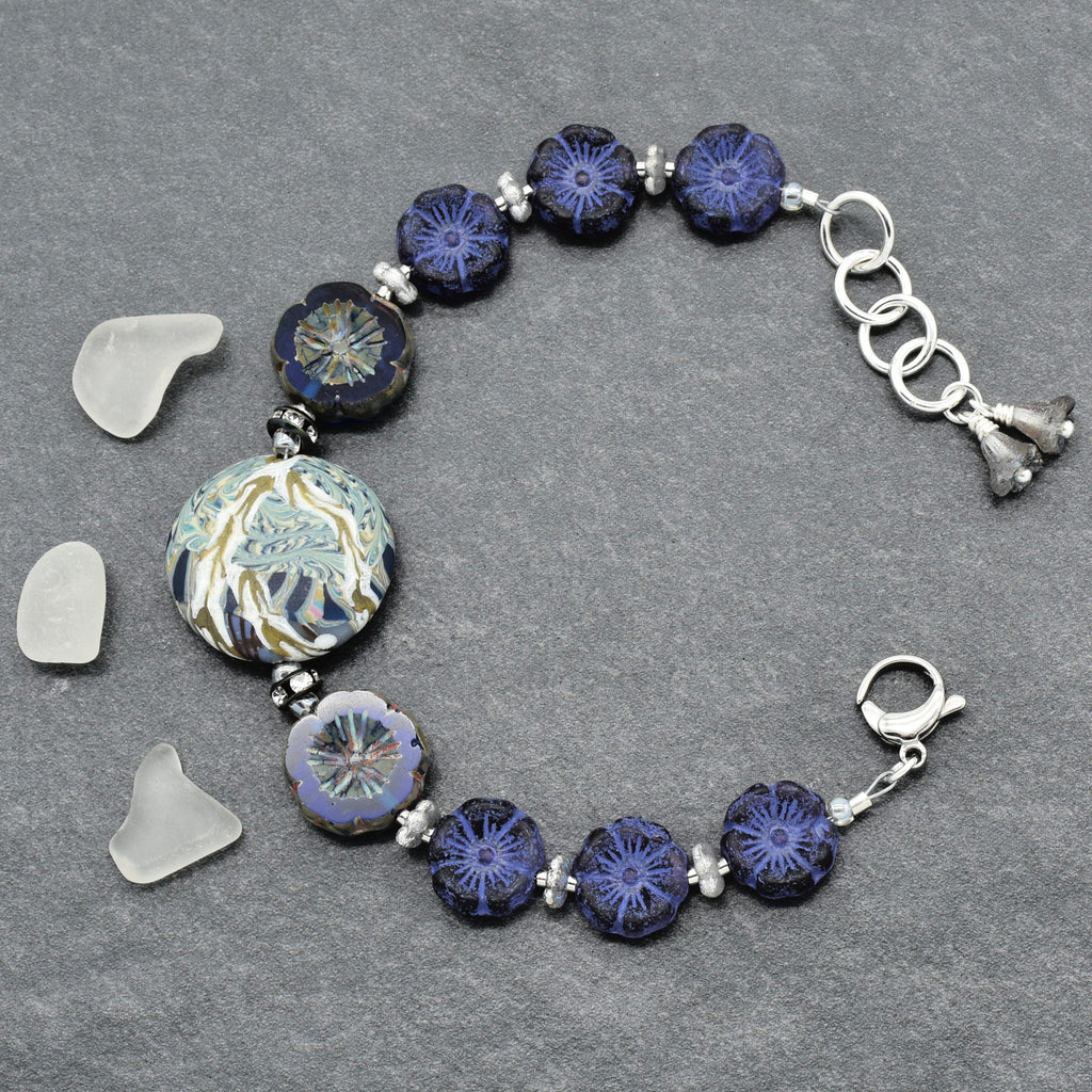 A handmade beaded bracelet featuring dark blue and indigo glass beads with intricate floral patterns, a large round patterned bead, and silver-toned metal links and charms, displayed on a textured gray background with three small sea glass pieces.