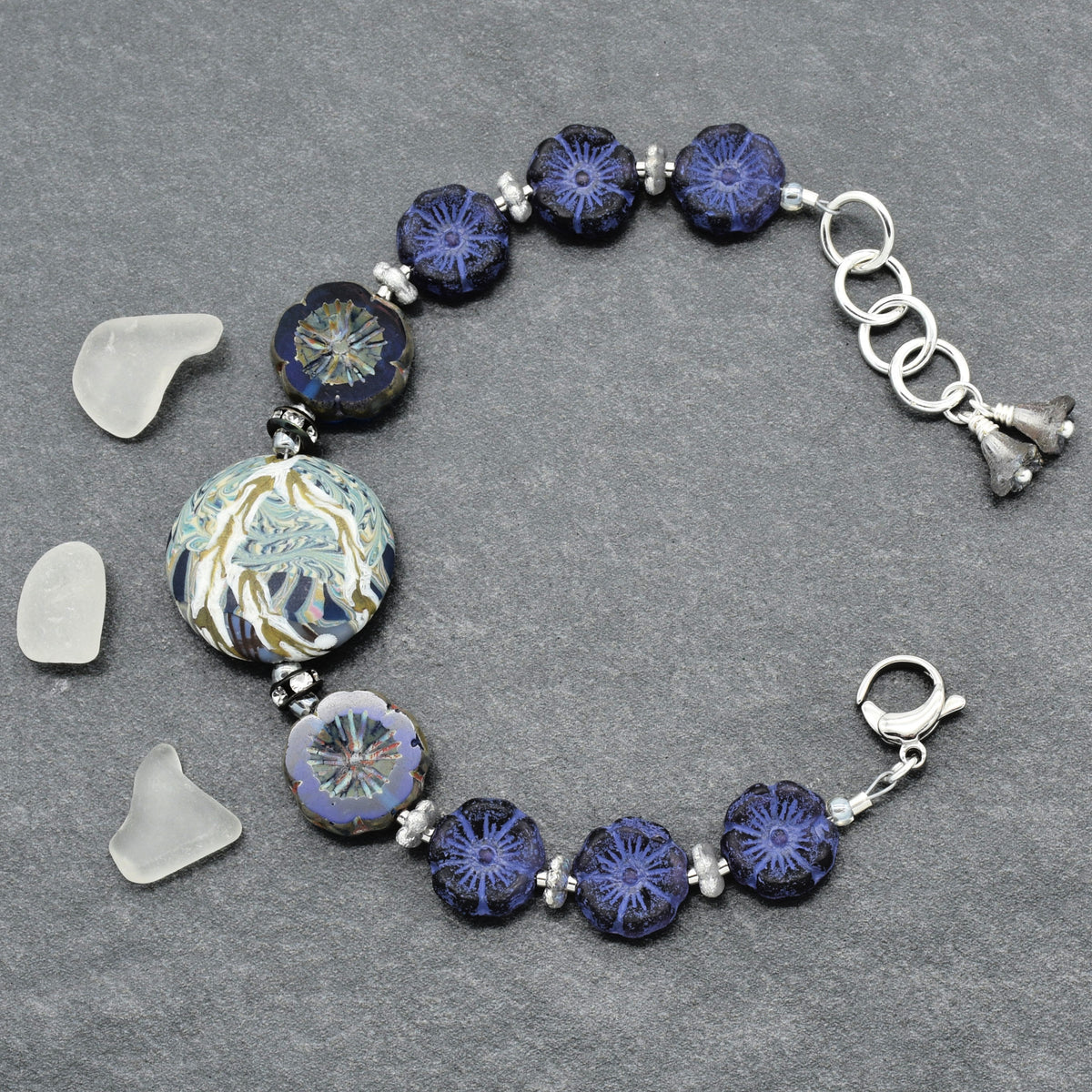 A handmade beaded bracelet featuring dark blue and indigo glass beads with intricate floral patterns, a large round patterned bead, and silver-toned metal links and charms, displayed on a textured gray background with three small sea glass pieces.