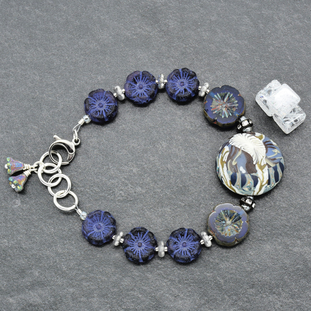A handmade glass bead bracelet featuring deep blue and indigo flower-shaped beads, a large round floral bead, and silver-toned metal rings and clasp, displayed on a textured gray background.