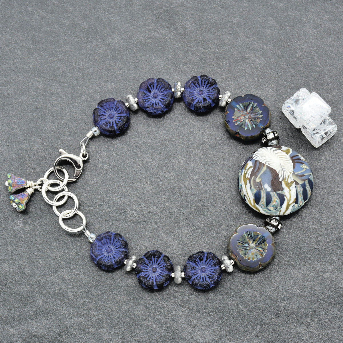 A handmade glass bead bracelet featuring deep blue and indigo flower-shaped beads, a large round floral bead, and silver-toned metal rings and clasp, displayed on a textured gray background.