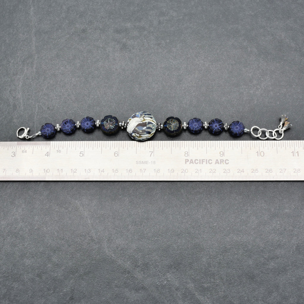 A beaded bracelet with dark blue faceted beads, a large floral patterned center bead, and silver-toned metal links, shown on a gray surface with a ruler for scale.