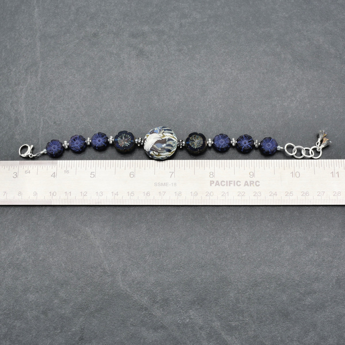 A beaded bracelet with dark blue faceted beads, a large floral patterned center bead, and silver-toned metal links, shown on a gray surface with a ruler for scale.