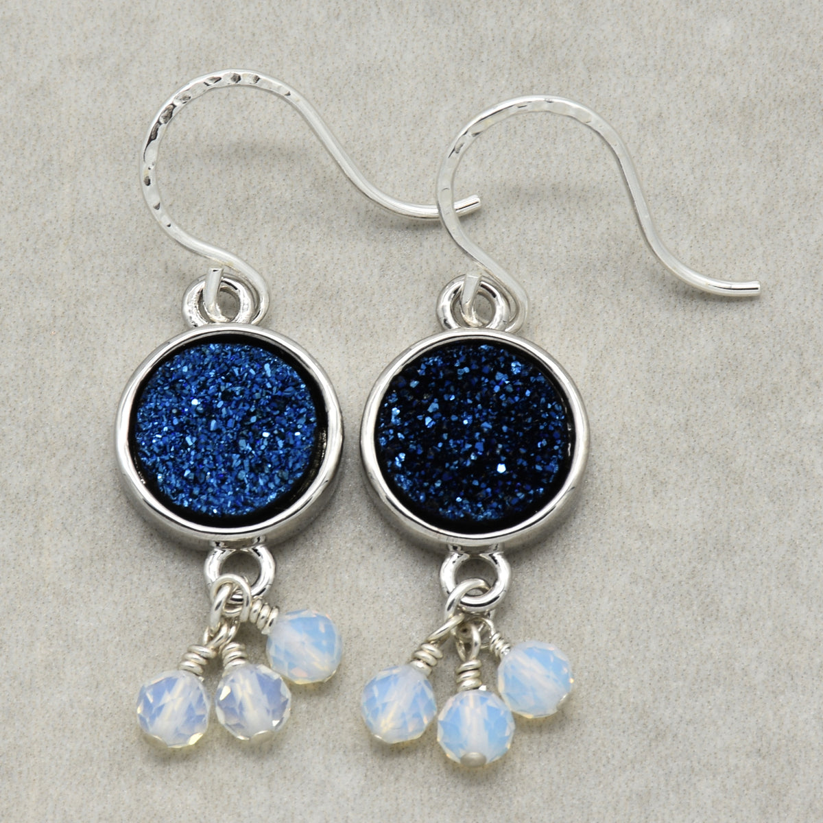 A pair of silver dangle earrings featuring round indigo crystal druzy links and three small white opalite beads hanging below each earring, displayed on a neutral textured gray background.