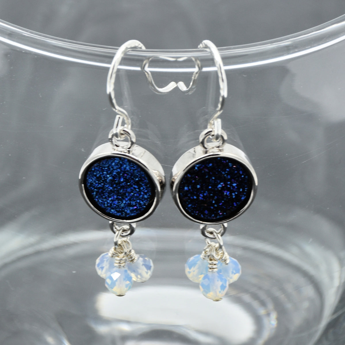 A pair of silver drop earrings with round indigo crystal druzy and dangling opalite faceted beads, displayed on a clear glass surface.