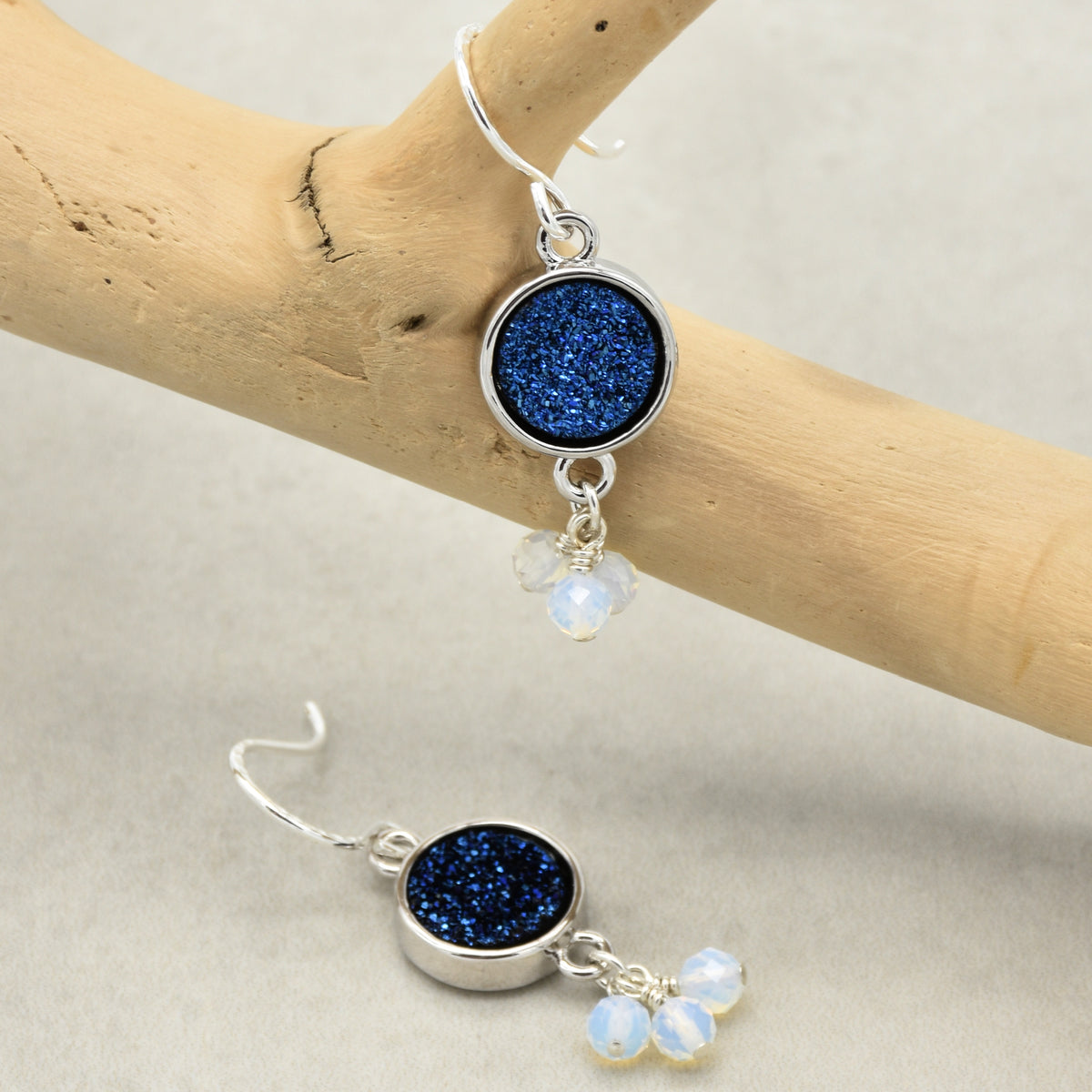 A pair of silver drop earrings with round indigo crystal druzy and small opalite bead charms, displayed on a light wood branch and neutral surface.