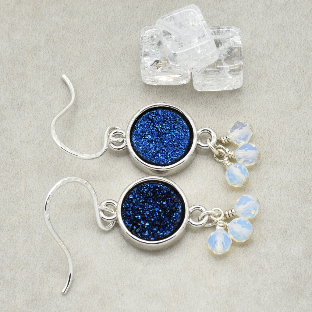 A pair of silver dangle earrings with round indigo crystal druzy links and small opalite bead drops, displayed on a light beige textured surface with clear crystal cubes in the background.