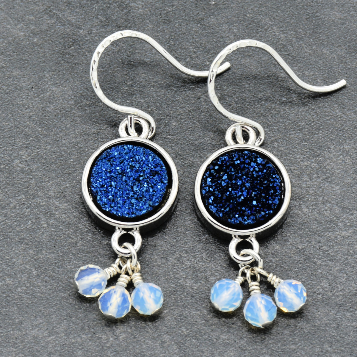 A pair of silver drop earrings featuring round indigo crystal druzy links and dangling opalite faceted beads, displayed on a dark gray textured background.