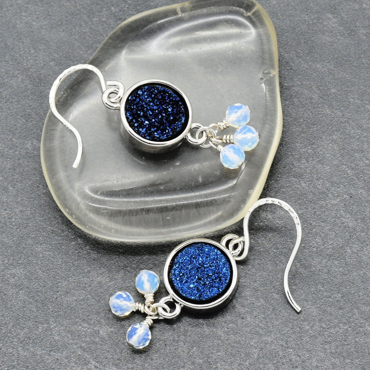 A pair of silver dangle earrings with round blue glitter centers and three small light blue faceted beads, displayed on a clear glass dish over a dark gray textured surface.