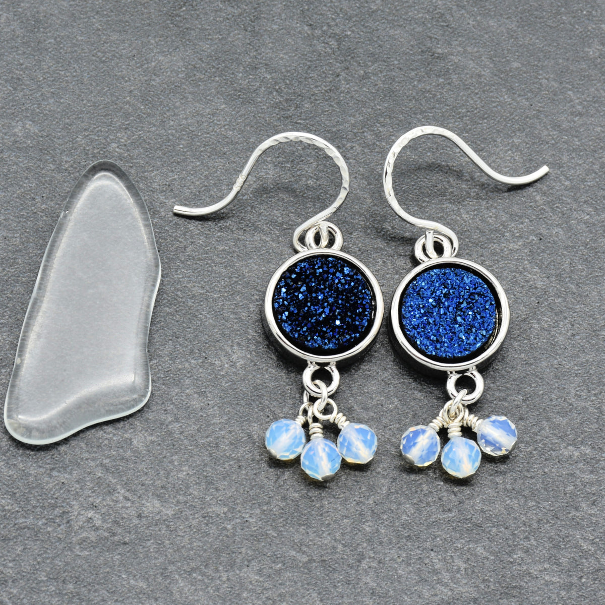 A pair of silver dangle earrings with round indigo crystal druzy links and small opalite bead drops, displayed on a dark gray textured surface next to a clear glass pebble.