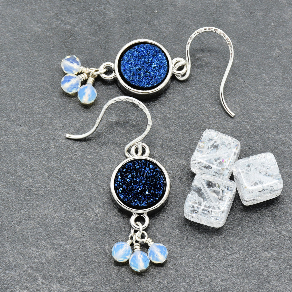 A pair of silver dangle earrings with round indigo crystal druzy links and small dangling opalite beads, displayed on a dark gray textured surface next to three clear ice cubes.