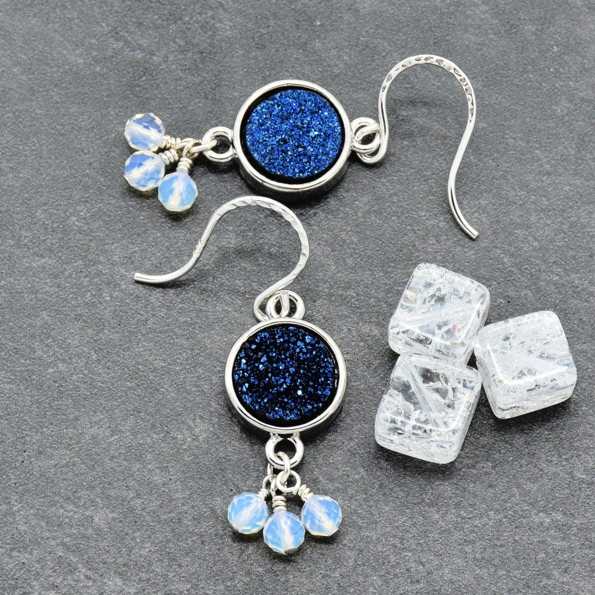 A pair of silver dangle earrings with round indigo crystal druzy links and small dangling opalite beads, displayed on a dark gray textured surface next to three clear ice cubes.