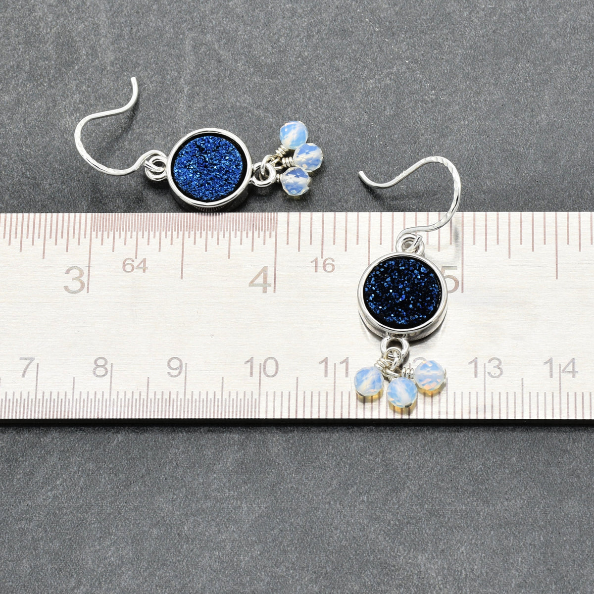 A pair of silver dangle earrings with round indigo crystal druzy links and small iridescent white beads, displayed above a metal ruler on a dark gray textured background.