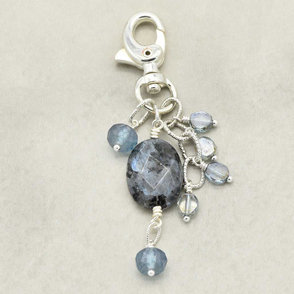A silver-toned keychain or bag charm featuring a faceted oval dark gray stone and several small round light blue beads, shown on a neutral beige background.