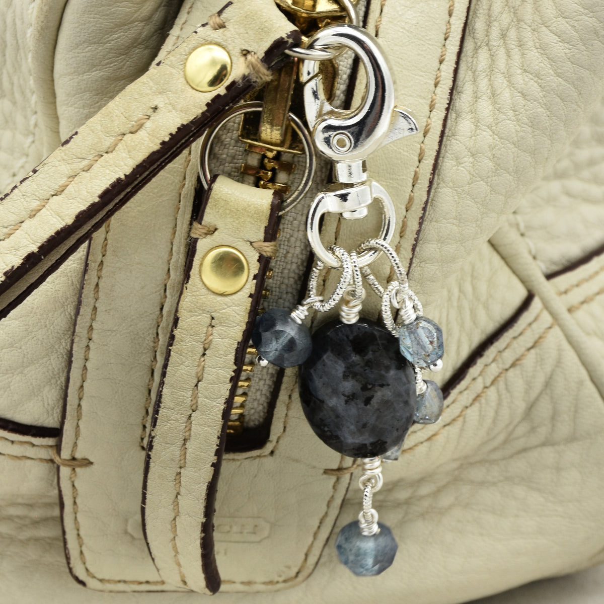 Close-up of a handbag zipper pull with a silver-tone clasp and charm featuring a large dark gray stone and several small blue faceted beads, attached to a light beige leather bag.