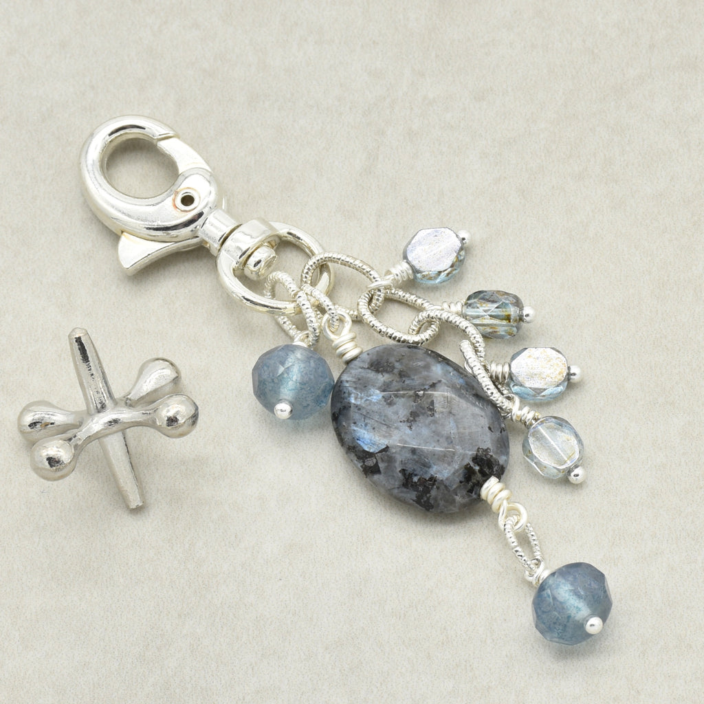 A silver lobster clasp keychain charm featuring a large oval dark gray labradorite stone and several dangling faceted beads in light blue and clear, shown on a neutral beige background.