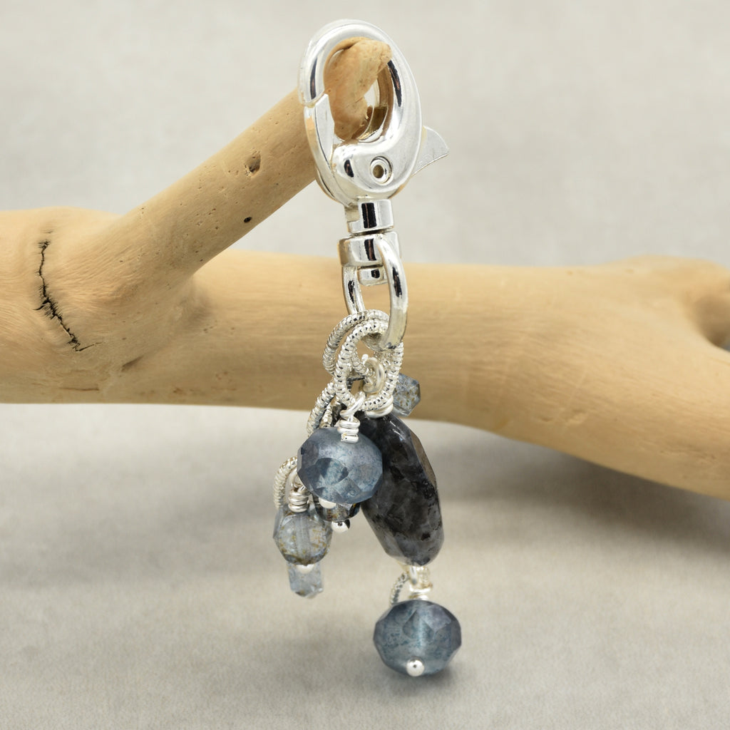 A silver-toned lobster clasp keychain or bag charm with dangling faceted beads in light blue, dark blue, and gray, displayed on a wooden stand against a neutral gray background.